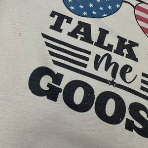 May include: A white t-shirt with black text that reads "TALK me GOOSE" with a star and three stripes above the text. The text is in a distressed font. There are two American flag sunglasses above the text.
