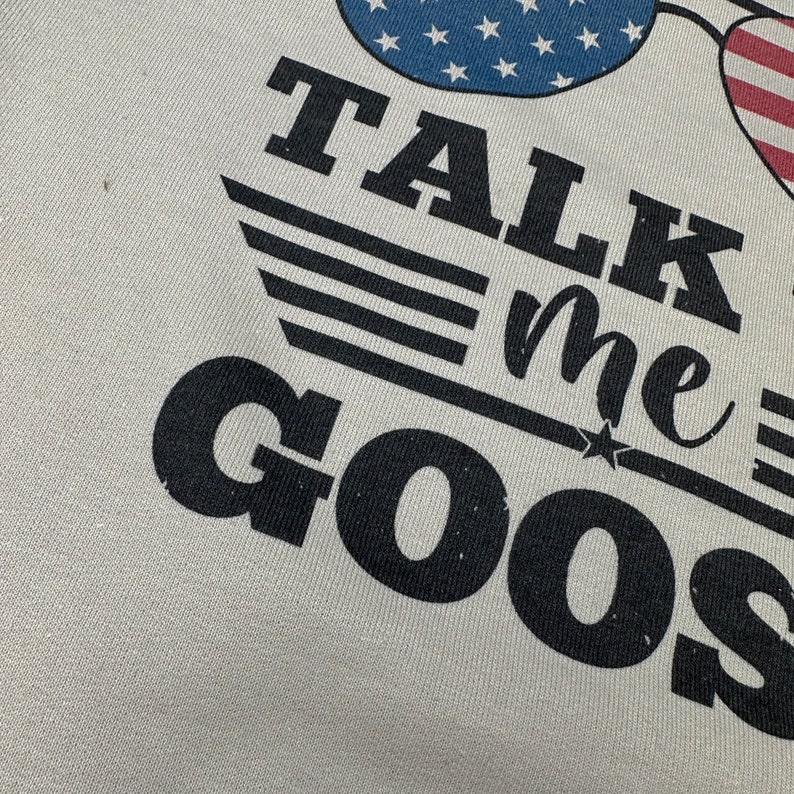 May include: A white t-shirt with black text that reads "TALK me GOOSE" with a star and three stripes above the text. The text is in a distressed font. There are two American flag sunglasses above the text.