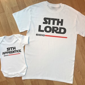 May include: White t-shirt with the text "Sith Lord" in black with a red lightsaber graphic. A white baby onesie with the text "Sith Apprentice" in black with a red lightsaber graphic.