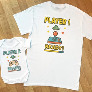 Op de afbeelding: Witte t-shirt en baby-romper set met bijpassende videogame-thema graphics. Het t-shirt zegt "PLAYER 1 READY!" en de romper zegt "PLAYER 2 READY!" met controller en ruimteschip illustraties. Perfect voor ouder en kind.