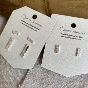 May include: Two pairs of silver bar earrings on white cards. The earrings have a textured surface. The cards have the text "OUTLINE JEWELLERY" and "Handmade in Recycled Silver www.outlinejewellery.com".