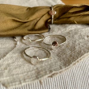 Peut inclure: Plusieurs bagues en argent, chacune ornée d'un détail ovale martelé. Les bagues sont disposées sur un tissu de lin beige, avec un tissu doré en arrière-plan. Le design des bagues est simple et minimaliste.