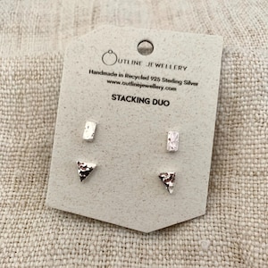 May include: A set of four silver stud earrings on a beige display card labelled OUTLINE JEWELLERY, Handmade in Recycled 925 Sterling Silver, STACKING DUO. The set includes two rectangular studs and two triangular studs with a textured, hammered finish.