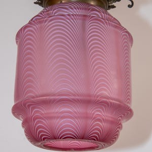 Könnte beinhalten: Vintage rosa Glas-Pendelleuchte mit dekorativer Messingfassung. Der Glasschirm hat ein wirbelndes weißes Muster. Die Leuchte ist an einer Kette aufgehängt und eignet sich für die Innenbeleuchtung.