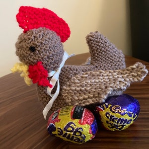 May include: A brown knitted chicken toy with a red comb and a white ribbon, sitting on three purple and gold foil-wrapped chocolate eggs.