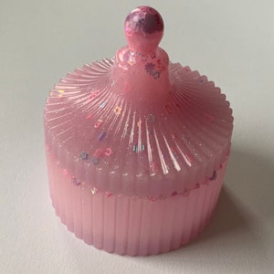 May include: A pink resin trinket box with a domed lid. The lid is decorated with glitter and small stars. The box has a ribbed design.