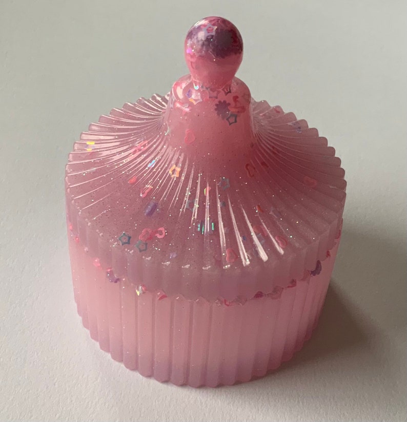 May include: A pink resin trinket box with a domed lid. The lid is decorated with glitter and small stars. The box has a ribbed design.