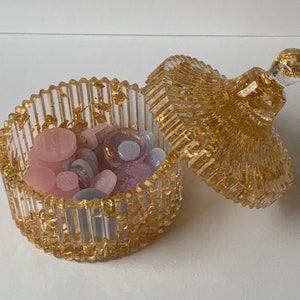 May include: A clear glass trinket box with a gold lid and gold flecks throughout. The box is filled with small pink, purple, and white buttons.