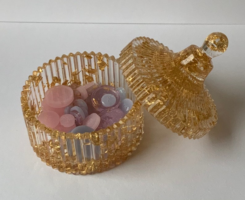 May include: A clear glass trinket box with a gold lid and gold flecks throughout. The box is filled with small pink, purple, and white buttons.