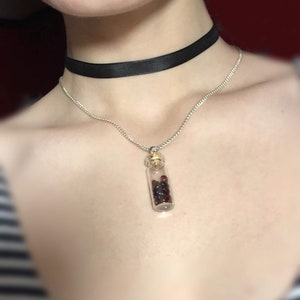 May include: A silver chain necklace with a small glass vial pendant filled with dark red beads. The necklace is worn over a black choker.