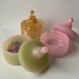 May include: Three decorative trinket boxes with lids. One box is green and pink with a ribbed design. Another box is pink with a ribbed design. The third box is gold with a ribbed design.