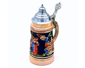 May include: A small, ceramic beer stein with a blue and white floral design and a silver lid. The stein features a scene of people in traditional German clothing.