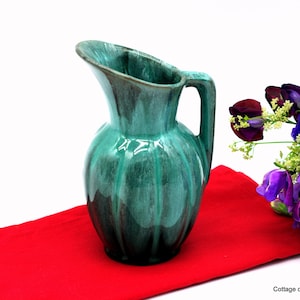 May include: A green ceramic jug with a textured glaze. The jug has a wide mouth and a handle. It is sitting on a red cloth.