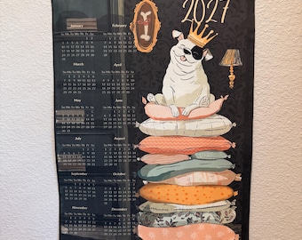 Può includere: Un calendario da parete 2027 stampato su tessuto con listelli di legno per appenderlo. Il design raffigura un cane bianco con una corona dorata, seduto su una pila di cuscini colorati. Sono visibili un'illustrazione di un osso e una lampada.