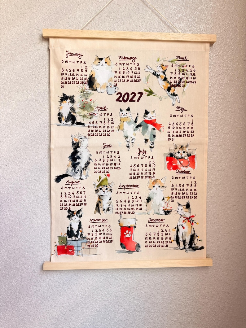 May include: A 2027 wall calendar printed on fabric with wooden hanging rods. The calendar features twelve months, each illustrated with a different cat in various poses and settings, including seasonal themes like a Christmas stocking and gifts.