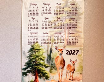 Op de afbeelding: Een wandkalender voor 2027, gedrukt op stof met houten ophangstokken. Het ontwerp bevat een aquarelillustratie van twee herten in een bosrijke omgeving met dennenbomen. De kalender toont alle twaalf maanden met datums in zwarte tekst.