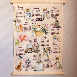 May include: A 2027 wall calendar printed on light-colored fabric with wooden hanging rods. The calendar features monthly grids and watercolor illustrations of various cats, kittens, and floral arrangements for each month of the year.