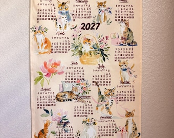 May include: A 2027 wall calendar printed on light-colored fabric with wooden hanging rods. The calendar features monthly grids and watercolor illustrations of various cats, kittens, and floral arrangements for each month of the year.