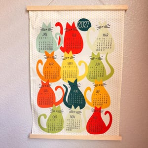 May include: A 2027 wall calendar printed on fabric with a light yellow polka dot background. The calendar features twelve colorful cat-shaped illustrations, each representing a month of the year with its corresponding dates and days of the week.
