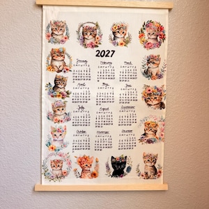 May include: A 2027 wall calendar printed on a rectangular fabric sheet with wooden hanging rods at the top and bottom. The calendar features twelve monthly grids, each decorated with a watercolor-style illustration of a kitten wearing a floral crown.