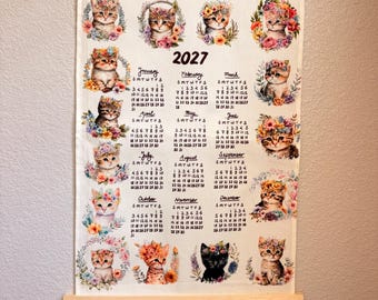 May include: A 2027 wall calendar printed on a rectangular fabric sheet with wooden hanging rods at the top and bottom. The calendar features twelve monthly grids, each decorated with a watercolor-style illustration of a kitten wearing a floral crown.