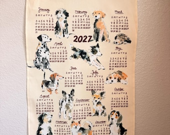 Può includere: Un calendario da parete in tessuto color crema per l'anno 2022, sospeso tramite binari in legno e uno spago. Il calendario presenta illustrazioni in stile acquerello di varie razze canine accanto a griglie mensili con le relative date.