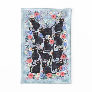 May include: A rectangular tea towel featuring a 2027 calendar design. The print displays twelve black cats, each representing a month, surrounded by a watercolor-style floral pattern with red, pink, and blue flowers on a light blue background.