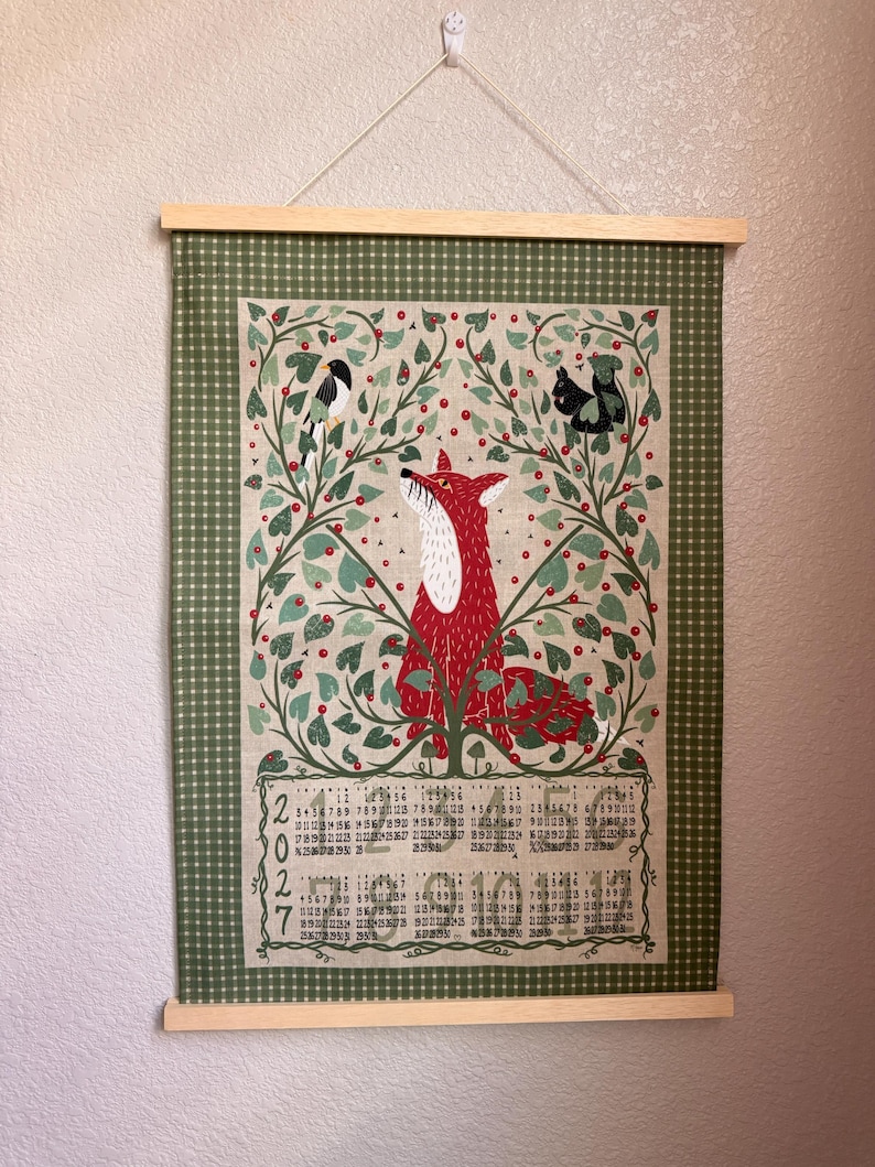 Op de afbeelding: Een wandkalender voor 2027, gedrukt op stof met een houten hanger. Het ontwerp toont een centrale rode vos omringd door groen gebladerte, rode bessen, een vogel en een eekhoorn. Het kalenderrooster staat onderaan in zwarte tekst.