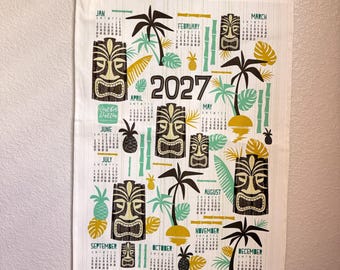 Puede incluir: Un calendario de pared para 2027 impreso en tela blanca con un soporte de madera en la parte superior e inferior. El diseño presenta máscaras tiki marrones, palmeras verdes, soles amarillos y piñas. El texto incluye el año 2027 y los meses.