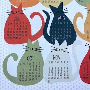 May include: A white fabric calendar featuring colorful cat-shaped silhouettes for each month. Each cat displays the month name, such as JUL, AUG, OCT, and NOV, along with the corresponding calendar dates in black text. The background has a light polka dot pattern.