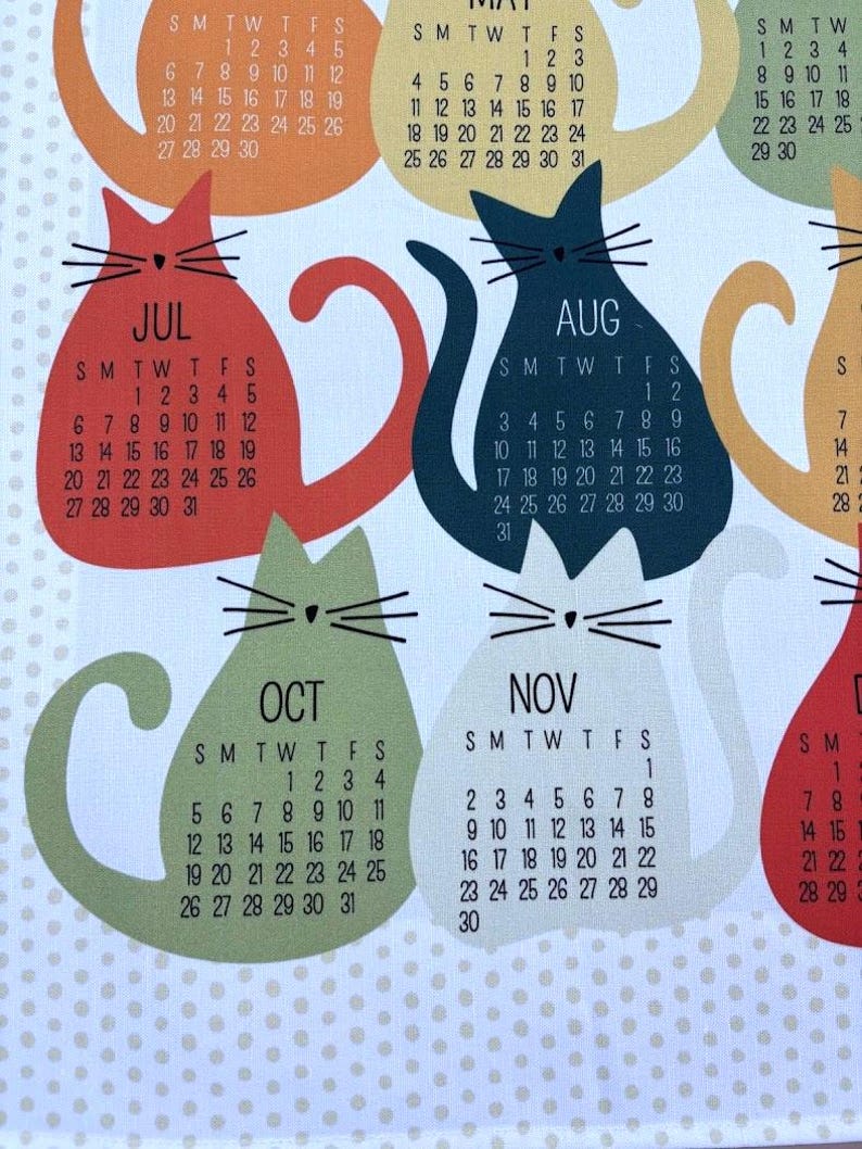 May include: A white fabric calendar featuring colorful cat-shaped silhouettes for each month. Each cat displays the month name, such as JUL, AUG, OCT, and NOV, along with the corresponding calendar dates in black text. The background has a light polka dot pattern.
