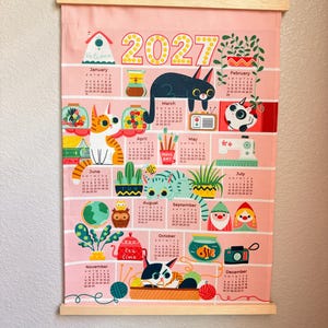 May include: A 2027 wall calendar printed on pink fabric with a light wood hanging frame. The calendar features twelve monthly grids decorated with colorful illustrations of cats, plants, household items, and whimsical characters in a grid layout.