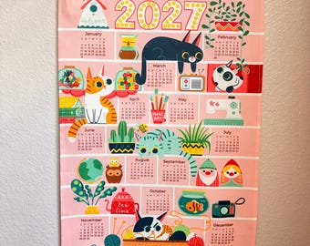 May include: A 2027 wall calendar printed on pink fabric with a light wood hanging frame. The calendar features twelve monthly grids decorated with colorful illustrations of cats, plants, household items, and whimsical characters in a grid layout.