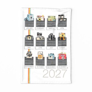 May include: A white tea towel featuring a 2027 calendar with a vertical rainbow stripe. Each month is represented by a unique illustration of a vintage camera in various colors, including blue, red, yellow, and black, positioned above the calendar grid.