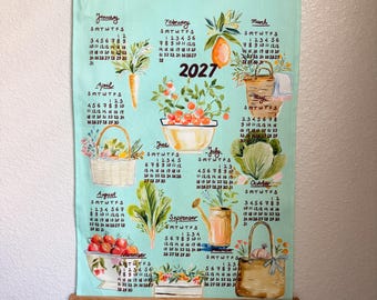 Puede incluir: Un calendario de pared para 2027 impreso en tela verde claro con varillas de madera para colgar. El calendario presenta ilustraciones en acuarela de productos de jardín, incluyendo zanahorias, limones, cestas, tomates y una regadera.