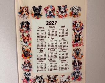 Può includere: Un calendario da parete 2027 stampato su tessuto color crema, sospeso tramite due barre di legno orizzontali e uno spago marrone. Il calendario presenta una griglia centrale dei mesi circondata da illustrazioni ad acquerello di varie razze canine.