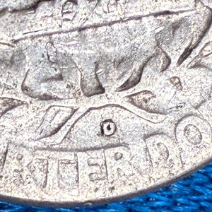May include: A silver-toned metallic coin featuring a raised relief design of a figure with a bow and arrow. The surface shows visible wear and texture. The letters "ER DO" are partially visible along the bottom edge, with a small circular mark.