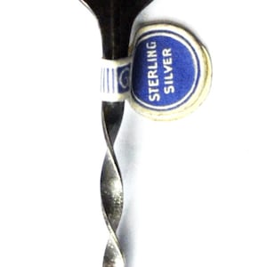 May include: A silver spoon with a twisted handle and a circular design on the end. The design features the words "World's Highest Peak" and a mountain range. The spoon is marked "Sterling Silver".