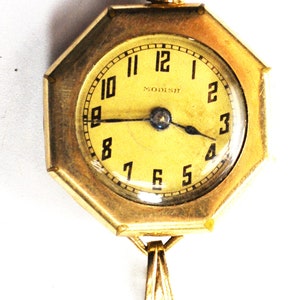 May include: A gold-toned octagonal pendant watch with a white face and black Roman numeral hour markers. The watch face also has black hands and the word "MODISH" printed in black.