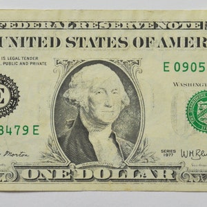 May include: A one dollar bill with a portrait of George Washington. The bill is green and white with the text "THE UNITED STATES OF AMERICA" and "ONE DOLLAR" printed on it. The serial number is E 09058479 E.