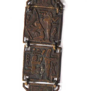 May include: A gold-toned metal pendant with an Egyptian theme. The pendant features four rectangular sections with raised images of Egyptian gods and goddesses. The bottom section is a round coin with a raised image of a woman's face.