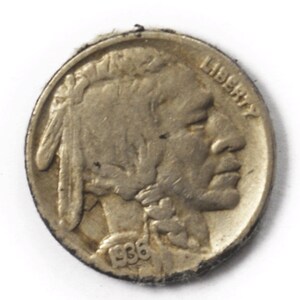 May include: A silver-colored US Buffalo nickel coin with the year 1936 inscribed on the bottom. The coin features a profile of a Native American with the word "LIBERTY" inscribed on the right side.