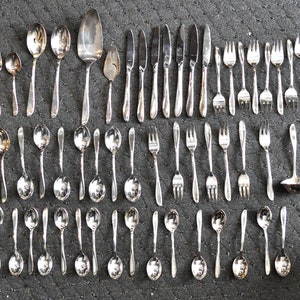 May include: A set of silverware, including forks, spoons, and knives, arranged in rows on a gray surface. The silverware is made of polished metal and has a traditional design.