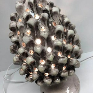 Puede incluir: Una lámpara de cerámica con forma de piña con un cable blanco. La lámpara tiene un esmalte marrón claro con una base blanca. La lámpara está encendida y la luz brilla a través de los agujeros del cono.