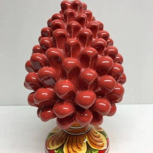 Puede incluir: Una escultura de piña de pino de cerámica roja con una base pintada. La base presenta un diseño floral con colores amarillos, naranjas y verdes. El texto "Ceramiche Siciliane" está escrito en la base.
