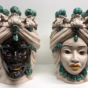 Puede incluir: Dos cabezas de maceta de cerámica con detalles verdes y blancos. Las cabezas llevan turbantes y tienen joyas elaboradas. Una cabeza es marrón y la otra es blanca.