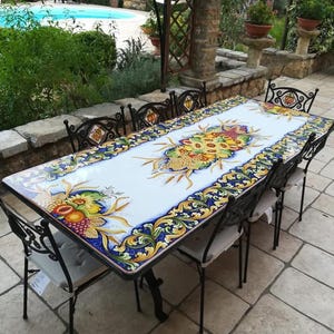 May include: A rectangular dining table with a white ceramic top featuring a vibrant, hand-painted border of yellow, blue, and green floral and fruit motifs. The table is accompanied by six black wrought iron chairs with light-colored seat cushions.