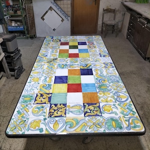 Può includere: Un tavolo rettangolare con un piano composto da numerose piastrelle di ceramica quadrate. Le piastrelle presentano un mix di colori solidi tra cui rosso, blu, giallo, verde e bianco, insieme a intricati motivi floreali dipinti a mano.