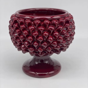 Puede incluir: Un macetero de cerámica rojo oscuro con forma de piña, con una abertura redonda ancha y una base de pedestal.
