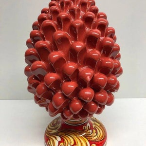 Puede incluir: Un gran cono de pino de cerámica roja con una superficie texturizada detallada. La base del cono está decorada con un patrón floral colorido y las palabras "Ceramiche Siciliane Caltagirone".